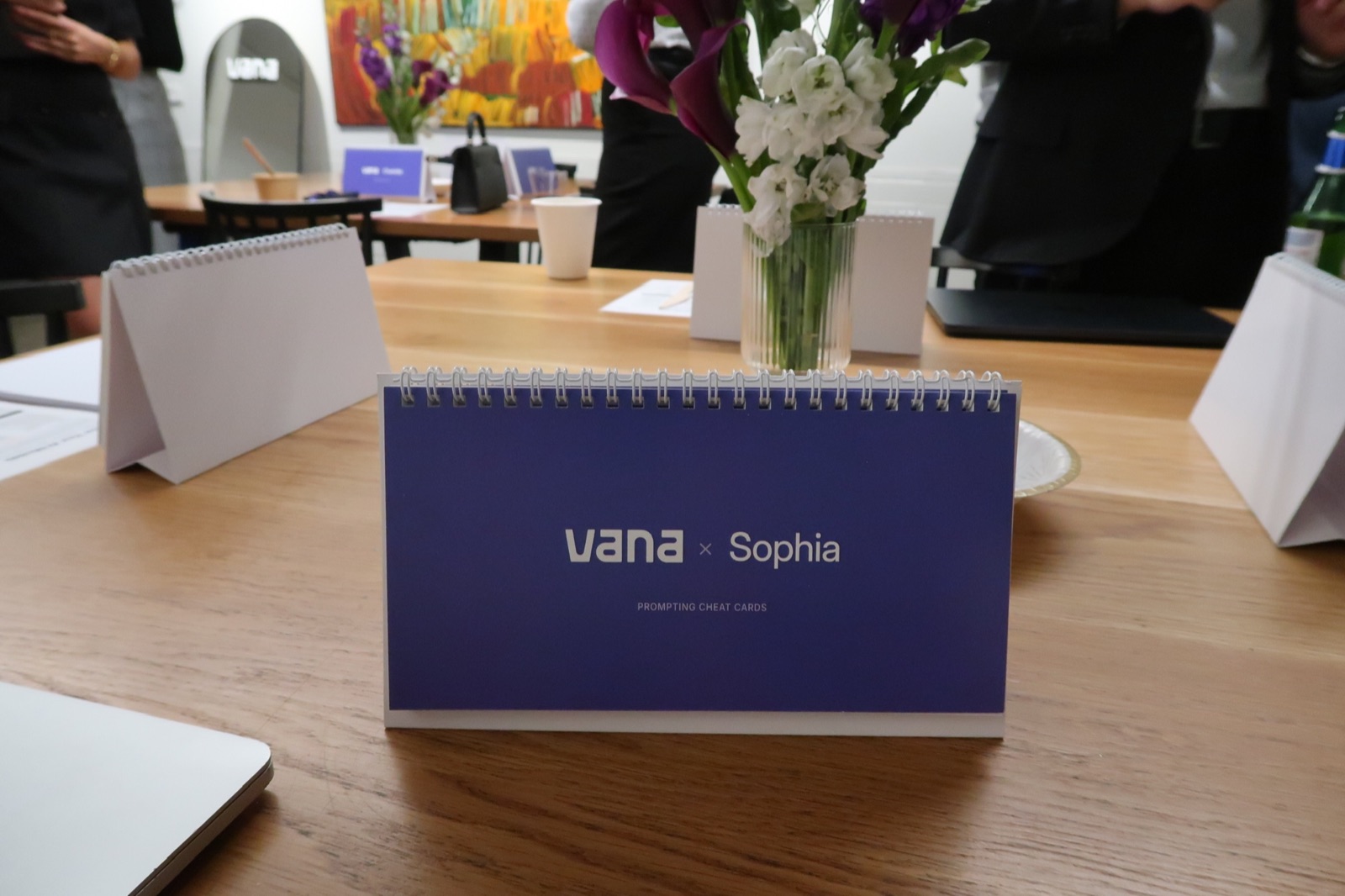 A personalised Vana prompting cheat card standing on a table beside flowers and laptops